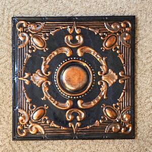 May include: Square, ornate ceiling tile with a black background and copper-colored accents. The design features a central circle, oval shapes, and scrolling patterns. The tile has a raised, textured appearance.