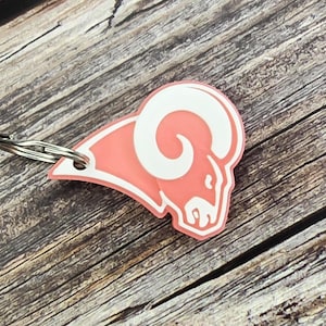 May include: A pink keychain with a white ram's head logo. The keychain is made of plastic and has a silver metal ring.