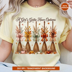 May include: A pale yellow t-shirt featuring the text A Girl's Gotta Have Options above five illustrated broomsticks. Each broom is decorated with autumn-themed elements including colorful bows, dried wheat, fall leaves, and small pumpkins.