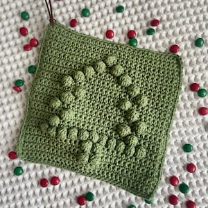 Może przedstawiać: Kwadratowy, zielony, szydełkowy podkładka pod garnek z wypukłym, teksturowanym wzorem w kształcie drzewa. Brązowa skórzana pętelka jest przymocowana do jednego rogu. Czerwone i zielone cukierki są rozrzucone wokół.