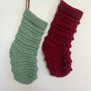 May include: Two handmade Christmas stockings. One is sage green, the other is deep red. Both are crocheted with a textured stitch and have a folded-over cuff. Each stocking has a brown leather loop for hanging.