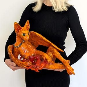 May include: A ceramic sculpture of a winged creature in shades of orange and red. The creature is reclining, with a necklace around its neck. The sculpture has a detailed texture and is held by a person wearing a black dress.