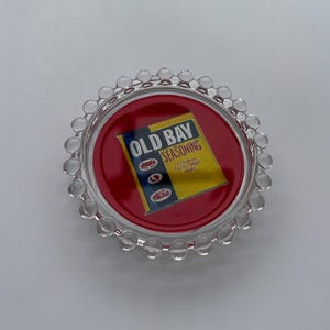 May include: A clear, round coaster with a red center featuring the "Old Bay Seasoning" logo in yellow and blue. The coaster has a clear, beaded edge.