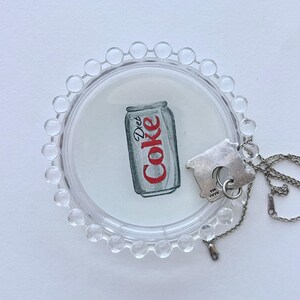 May include: A clear, round, beaded tray holds a watercolor illustration of a Diet Coke can. A silver-toned house-shaped pendant hangs on a delicate chain, resting near the tray.