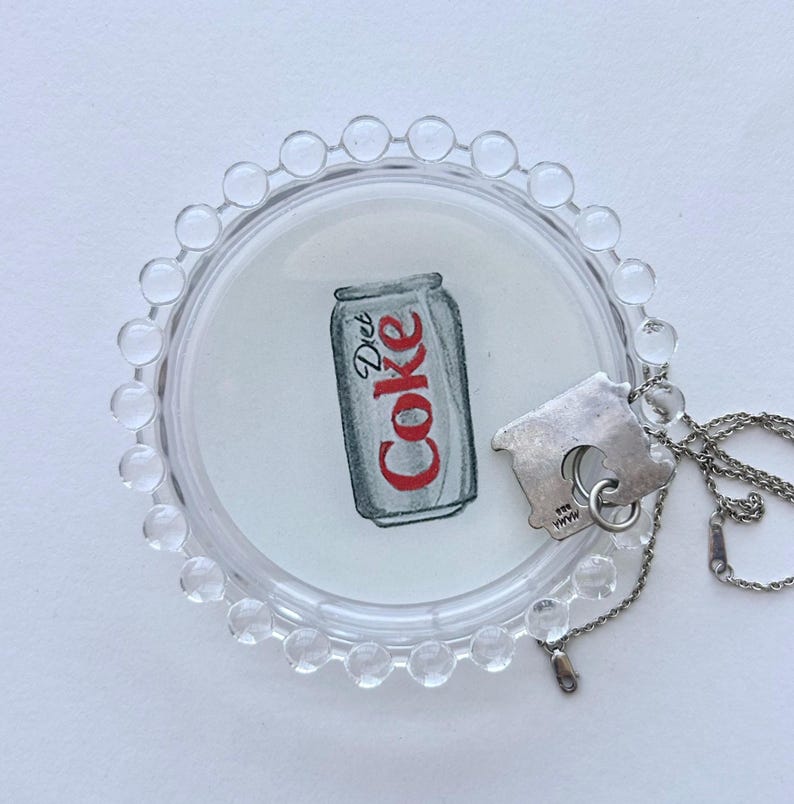 May include: A clear, round, beaded tray holds a watercolor illustration of a Diet Coke can. A silver-toned house-shaped pendant hangs on a delicate chain, resting near the tray.