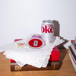 May include: A Diet Coke can with red lettering sits atop a stack of books and a red box. A small Coke pin is displayed in a clear oval frame with a red background. A small paper sign that says "Wear Your Snacks" is also present.