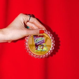 May include: A circular coaster with a clear, beaded edge featuring a miniature graphic of a Cheetos Flamin' Hot snack bag in the center. The snack bag graphic displays the Cheetos logo, the text Flamin' Hot, and the cartoon cheetah mascot.
