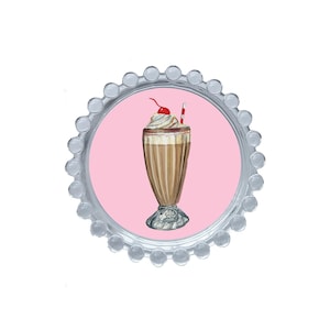 Puede incluir: Una tapa de botella plateada con un centro rosa que presenta una ilustración de batido. El batido es marrón con crema batida, una cereza y una pajita roja. La tapa tiene un borde festoneado.