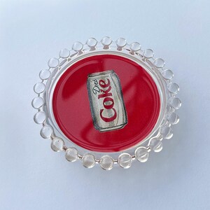 May include: A round, clear acrylic coaster with a red center featuring a Diet Coke can illustration. The coaster has a decorative beaded edge. The Diet Coke can is light gray with red and white text.
