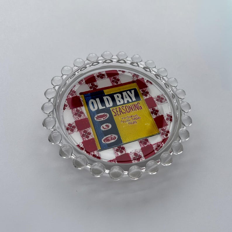 May include: A clear glass dish with a scalloped edge. The dish features a red and white checkered pattern and the words "OLD BAY SEASONING" on a yellow background. The dish is designed for serving.