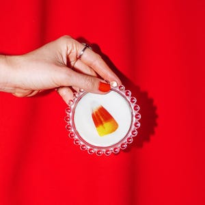 May include: A round, clear glass coaster with a beaded edge, featuring a single piece of orange, yellow, and white candy corn centered on its white surface. The coaster is held against a solid, vibrant red background.