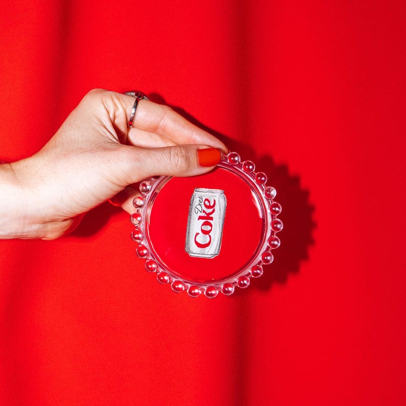 May include: A clear, round coaster with a red center and a Diet Coke can graphic. The coaster has a beaded edge and is held against a solid red background.