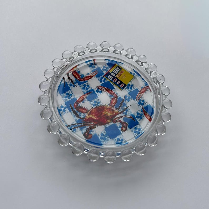 Puede incluir: Plato de cristal transparente con dise&ntilde;o de cangrejo y fondo a cuadros azules y blancos. El plato tiene un borde con cuentas y presenta el texto "Old Bay Seasoning".