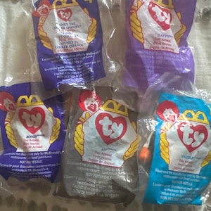 Può includere: Cinque giocattoli Ty Beanie Baby sigillati nella loro confezione originale. Le confezioni sono di vari colori, tra cui viola, blu e grigio, e presentano il logo a cuore Ty e nomi di personaggi.