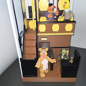 May include: A detailed diorama of The Muppet Show stage, featuring figures of Fozzie Bear, Kermit the Frog, Bert, and Ernie. The set includes a staircase, doors, and a sign that reads "EXIT". The base has a black oval with "The Muppet Show 25 Years" printed on it.