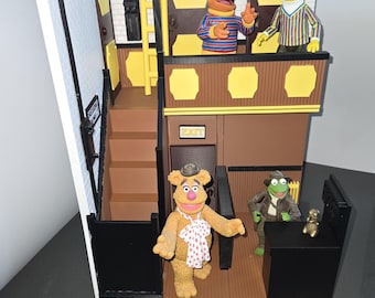 May include: A detailed diorama of The Muppet Show stage, featuring figures of Fozzie Bear, Kermit the Frog, Bert, and Ernie. The set includes a staircase, doors, and a sign that reads "EXIT". The base has a black oval with "The Muppet Show 25 Years" printed on it.