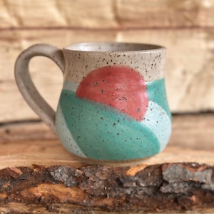 May include: A speckled ceramic mug with a rounded body and a curved handle. The exterior features a hand-painted design with a large red semi-circle shape above overlapping wavy bands of dark teal and light blue against a beige speckled base.