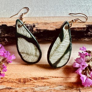May include: A pair of teardrop-shaped earrings featuring a dark green and cream-colored leaf pattern. The earrings are suspended from gold-toned metal hooks and display a smooth, glossy finish with intricate line details resembling botanical veins.