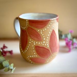 May include: A ceramic mug with a speckled tan base featuring a repeating pattern of terracotta-colored leaf shapes outlined by small, raised white dots. The interior of the mug is white, and it includes a curved handle on the left side.