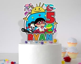 May include: A colorful cake topper featuring cartoon characters and the name "RYAN" in rainbow letters. The number "5" is prominently displayed, suggesting a birthday theme. The cake is white, with cupcakes in the background.