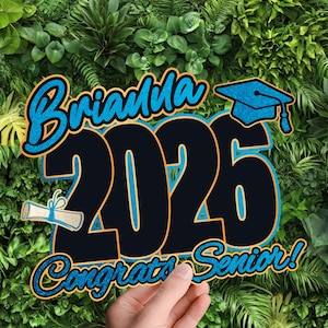 Op de afbeelding: Een afstudeerbord met de naam "Brianna" in blauw glitterscript boven de cijfers "2026" in zwart met een blauwe omlijning. De woorden "Congrats Senior!" staan eronder. Een afstudeerhoed en een diploma zijn ook afgebeeld.
