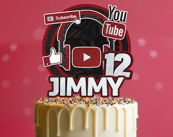 Puede incluir: Una tarta con glaseado blanco y chispas de colores, coronada con un adorno de tarta personalizado. El adorno presenta un tema de YouTube con las palabras "Suscríbete" y "You Tube", un pulgar hacia arriba, un botón de reproducción, el número "12" y el nombre "JIMMY".