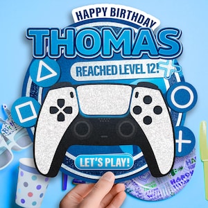 Puede incluir: Decoración de cumpleaños azul y blanca con un diseño de mando de videojuegos. El texto dice "HAPPY BIRTHDAY THOMAS REACHED LEVEL 12!" y "LET'S PLAY!". Suministros para fiestas, incluyendo gafas y una taza, son visibles en el fondo.
