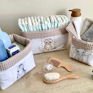Könnte beinhalten: Drei Stoffaufbewahrungskörbe mit beigem Waffelstrickrand und Tierillustrationen, gefüllt mit Windeln, einer Mustela-Schachtel und gefalteten Tüchern. Ein Holzkamm, eine weiche Holzbürste und ein Schnuller liegen davor.