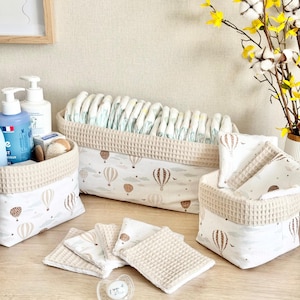 Op de afbeelding: Een set van drie stoffen opbergmanden en diverse vierkante doekjes met een beige wafelstof rand en witte katoenen stof bedrukt met bruine en beige heteluchtballonnen en wolken. Een doorzichtige plastic fopspeen ligt op de voorgrond.