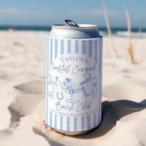 Op de afbeelding: Een blauw-witte blikjeskoeler met de tekst "Taylor's Coastal Cowgirl Bach Club Santa Rosa Beach, Florida" en een cartoonillustratie van een cowgirl in een martiniglas. De koeler staat op een zandstrand.