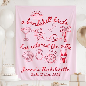May include: A pink fabric banner with red illustrations and text. The banner reads "a bombshell bride has entered the villa" with drawings of a martini, bikini, ring, and disco ball. The text "Jenna's Bachelorette Lake Tahoe 2025" is also included.