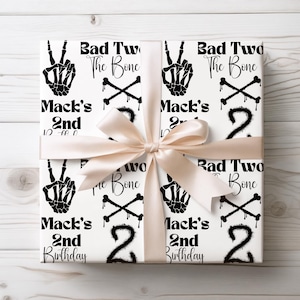 May include: White wrapping paper with a black and white skeleton hand peace sign, crossbones, and the text "Bad Two The Bone" and "Mack's 2nd Birthday".