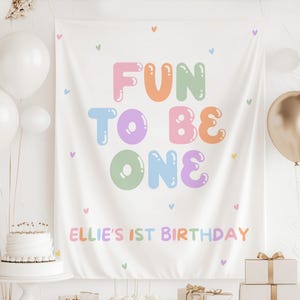 Op de afbeelding: Een witte stoffen banner met de tekst "FUN TO BE ONE" in kleurrijke bubbelletters. De banner bevat ook de tekst "ELLIE'S 1ST BIRTHDAY" en kleine hartvormen. Ballonnen en een taart staan op de achtergrond.