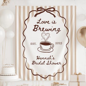 May include: A banner with the text "Love is Brewing" and "Hannah's Bridal Shower" in brown script. The banner features a coffee cup graphic and is surrounded by a brown decorative border. White and gold balloons and a cake are in the background.