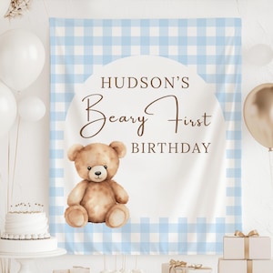 May include: A blue and white checked backdrop with a white circle displaying the text "Hudson's Beary First Birthday" and a teddy bear illustration. White and gold balloons and a cake are also visible.