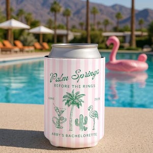 Op de afbeelding: Een roze en witte gestreepte blikjeskoeler met de tekst "Palm Springs Before the Rings" en illustraties van een palmboom, cactus en flamingo. De tekst "Abby's Bachelorette" is ook opgenomen. Een zwembad en een roze flamingo-drijver zijn op de achtergrond.