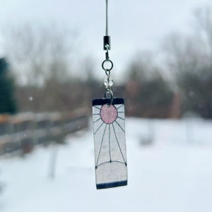 May include: A white and black charm with a pink sun design hanging from a silver chain. The charm is a popular anime character symbol.