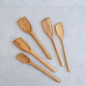 May include: A set of five light brown wooden cooking spoons with flat, angled edges and long, slender handles. Each spoon displays a distinct natural wood grain pattern, varying in size from a large, wide-headed spoon to a smaller, narrow-headed spoon.