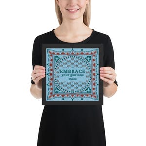 Puede incluir: Una impresión artística enmarcada con el texto "EMBRACE your glorious mess". La obra presenta un fondo azul claro con un diseño geométrico en rojo, verde azulado y blanco. El marco es negro.