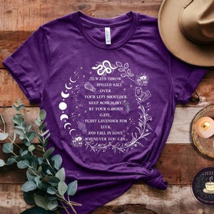May include: Purple t-shirt with a white graphic design featuring a moon phase, a snake, and floral elements. The text on the shirt reads "Always throw spilled salt over your left shoulder. Keep rosemary by your garden gate, plant lavender for luck, and fall in love whenever you can."