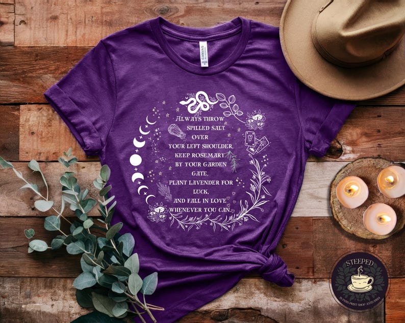 May include: Purple t-shirt with a white graphic design featuring a moon phase, a snake, and floral elements. The text on the shirt reads "Always throw spilled salt over your left shoulder. Keep rosemary by your garden gate, plant lavender for luck, and fall in love whenever you can."