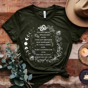 May include: A dark green t-shirt with a white graphic design featuring a moon phase, a plant, and text that reads "Always throw spilled salt over your left shoulder. Keep rosemary by your garden gate. Plant lavender for luck. And fall in love whenever you can."