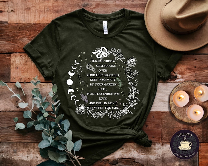 May include: A dark green t-shirt with a white graphic design featuring a moon phase, a plant, and text that reads "Always throw spilled salt over your left shoulder. Keep rosemary by your garden gate. Plant lavender for luck. And fall in love whenever you can."