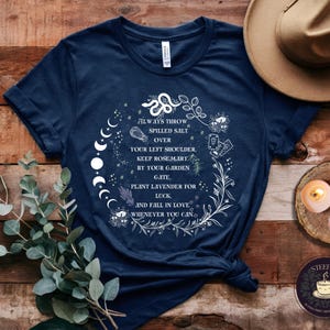 May include: A navy blue t-shirt with a white graphic design featuring a circle of moon phases, flowers, and the text "ALWAYS THROW SPILLED SALT OVER YOUR LEFT SHOULDER, KEEP ROSEMARY BY YOUR GARDEN GATE. PLANT LAVENDER FOR LUCK, AND FALL IN LOVE WHENEVER YOU CAN."