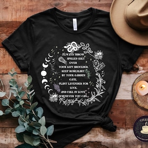 Puede incluir: Camiseta negra con un diseño gráfico blanco que presenta una fase lunar, flores y el texto "Siempre tira la sal derramada sobre tu hombro izquierdo. Mantén romero junto a la puerta de tu jardín, planta lavanda para la suerte y enamórate siempre que puedas."