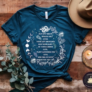 May include: Teal blue t-shirt with a white graphic design featuring a moon phase, a snake, and floral elements. The text on the shirt reads "Always throw spilled salt over your left shoulder, keep rosemary by your garden gate, plant lavender for luck, and fall in love whenever you can."