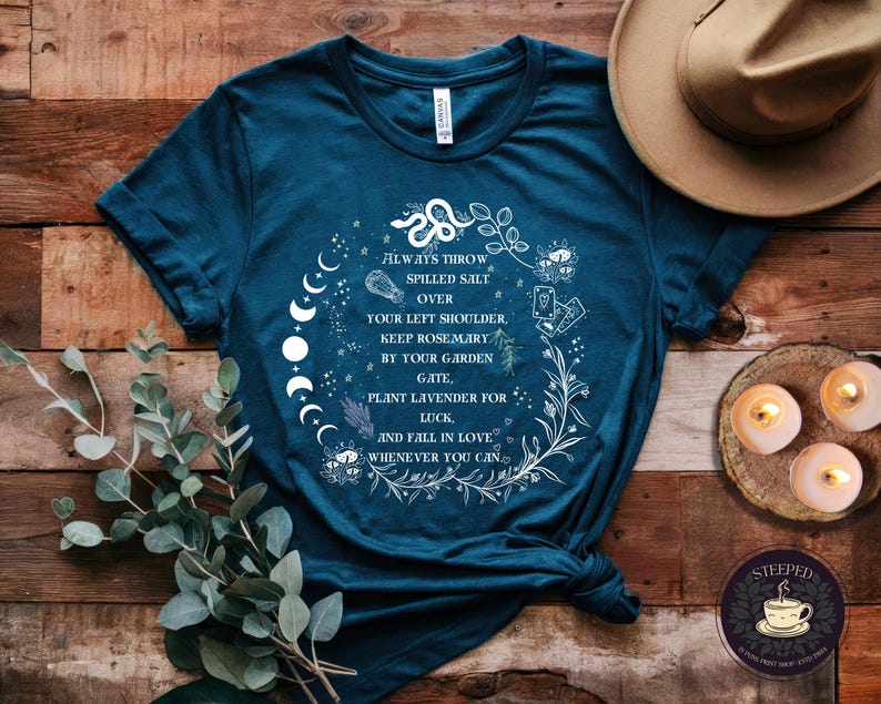 May include: Teal blue t-shirt with a white graphic design featuring a moon phase, a snake, and floral elements. The text on the shirt reads "Always throw spilled salt over your left shoulder, keep rosemary by your garden gate, plant lavender for luck, and fall in love whenever you can."