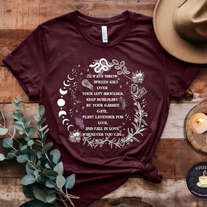 May include: A maroon t-shirt with a white graphic design featuring a moon phase, a snake, and floral elements. The text on the shirt reads "Always throw spilled salt over your left shoulder. Keep rosemary by your garden gate, plant lavender for luck, and fall in love whenever you can."