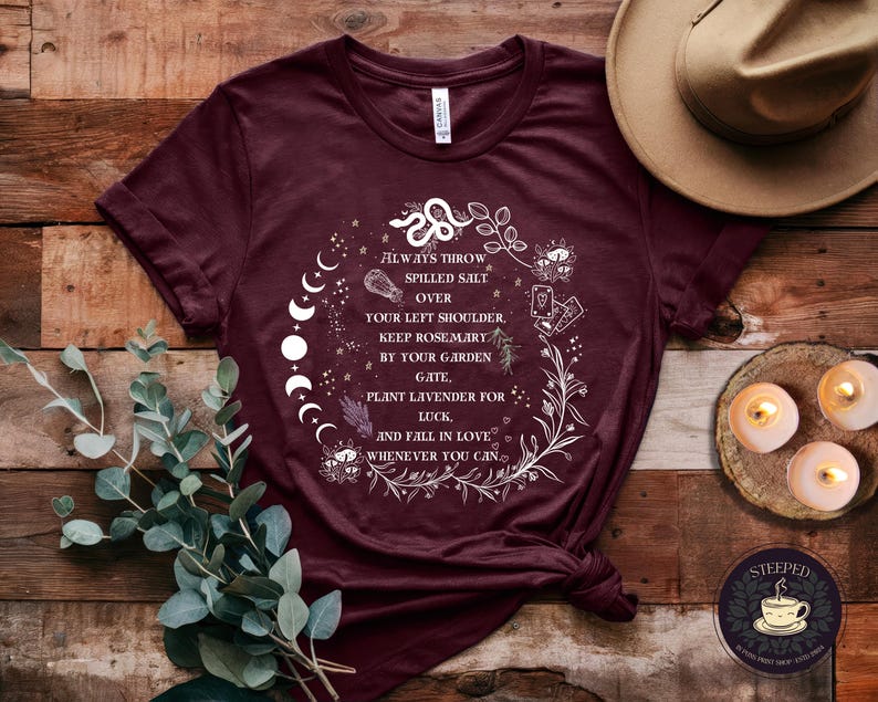 May include: A maroon t-shirt with a white graphic design featuring a moon phase, a snake, and floral elements. The text on the shirt reads "Always throw spilled salt over your left shoulder. Keep rosemary by your garden gate, plant lavender for luck, and fall in love whenever you can."