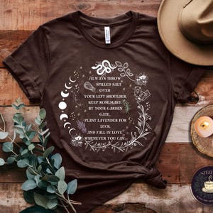 May include: A brown t-shirt with a white graphic design featuring a moon phase, a snake, and text that reads "Always throw spilled salt over your left shoulder. Keep rosemary by your garden gate, plant lavender for luck, and fall in love whenever you can."